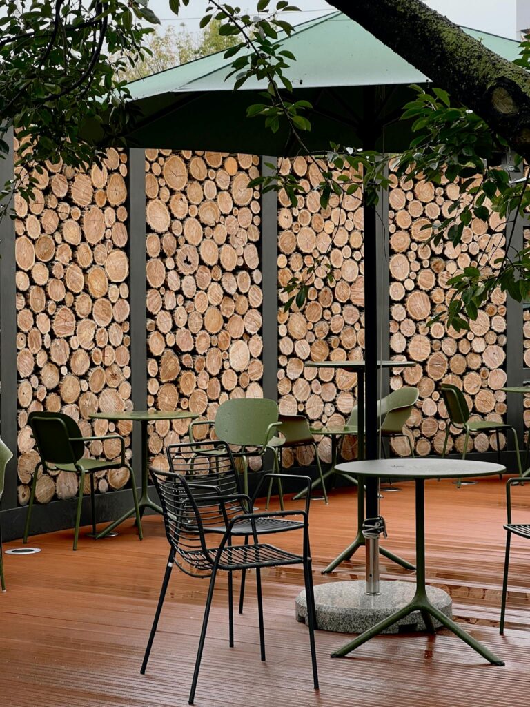 Cozy outdoor café setting with rustic wood stack walls, green tables, and chairs under an umbrella.